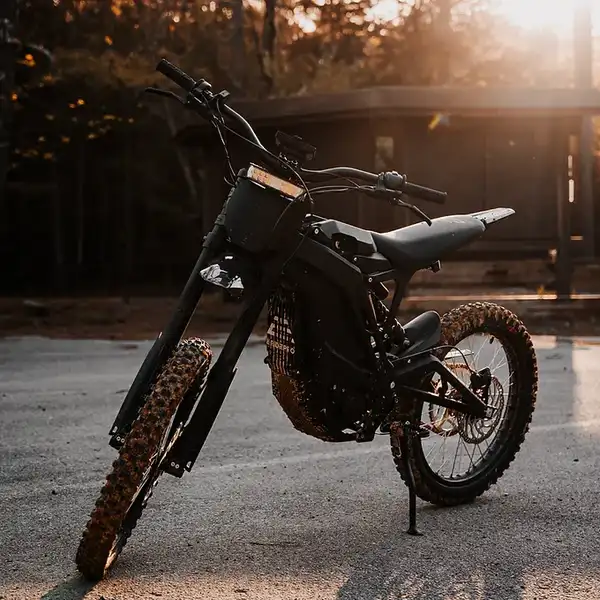 Black off-road motorcycle with thick knobby tires parked on a paved surface at sunset, leaning on its kickstand with a warm background glow.
