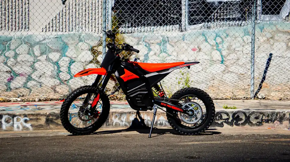 Orange dirt bike motorcycle with a black frame and orange body panels, parked on a street with a graffiti-covered wall behind it, on a side stand.