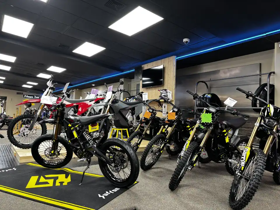 Showroom displaying multiple dirt bikes in a motorcycle shop, lined up on the floor under bright ceiling lights.